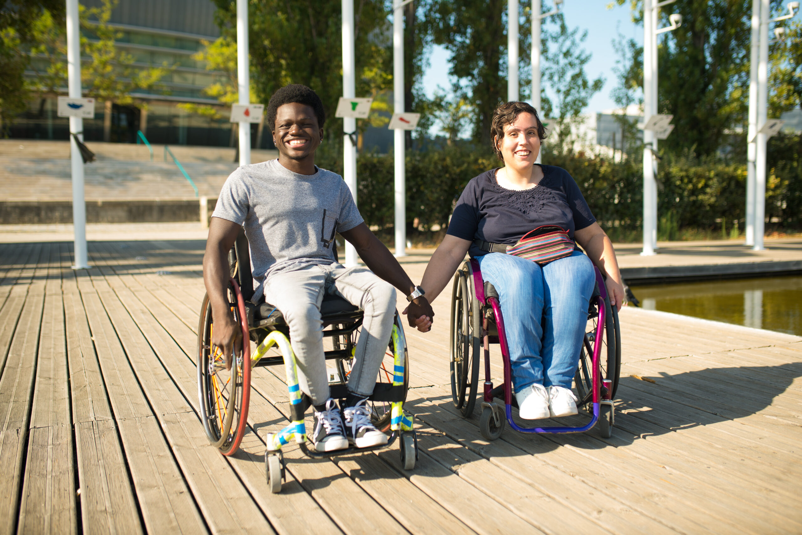 A man and a woman who both use wheelchairs sit together near the water, holding hands