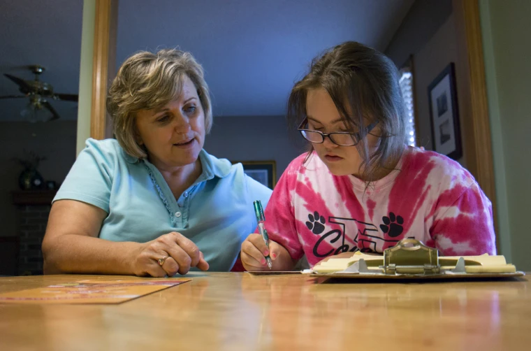 A caregiver sits beside a young woman as she writes on a clipboard at a table